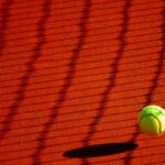 A vibrant tennis ball casting a shadow on a sunlit clay court with net shadows.